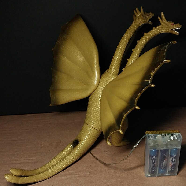 May include: A gold-coloured three-headed dragon toy figure with textured scales and large wings. The figure is connected by a thin wire to a clear plastic battery pack containing three blue cylindrical batteries, providing power to the creature.