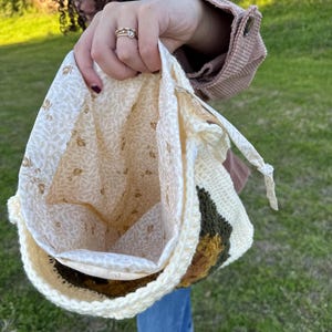 Puede incluir: Un bolso de ganchillo color crema con forro estampado floral. El bolso presenta un diseño geométrico en tonos marrón, verde y crema. El bolso se sostiene abierto, revelando el interior y el cierre de cordón.