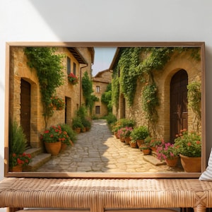 May include: A framed print showing a charming European street scene. The artwork features a cobbled path flanked by buildings, decorated with flowers and foliage. The print is displayed on a woven bench with books and sandals.