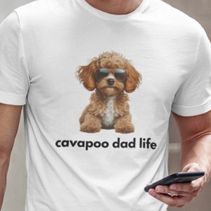 May include: White t-shirt featuring a brown Cavapoo dog wearing sunglasses. The text "cavapoo dad life" is printed below the dog. The shirt is paired with blue jeans.