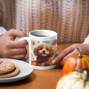 May include: White ceramic mug featuring a brown and white dog wearing a red scarf, printed against a backdrop of falling autumn leaves. The mug is filled with a brown liquid.