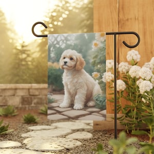 May include: A garden flag featuring a white and cream coloured cockapoo puppy sitting on a stone path with white flowers in the background.