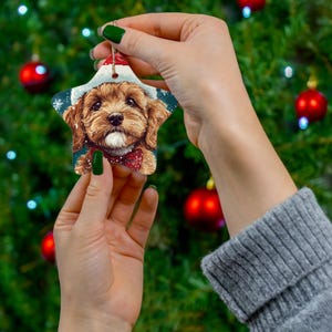 May include: A ceramic Christmas bauble shaped like a star featuring a brown and white dog wearing a Santa hat. The bauble has a blue background with white snowflakes.