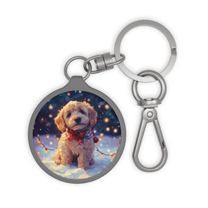 Può includere: Un portachiavi argentato con un ciondolo rotondo che presenta un piccolo cane marrone chiaro che indossa un fiocco rosso e una ghirlanda di luci. Il cane è seduto sulla neve con uno sfondo blu.
