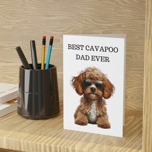May include: A white greeting card featuring the text "BEST CAVAPOO DAD EVER" and a cartoon Cavapoo dog wearing sunglasses. The card is on a wooden shelf, next to a black pen holder with pens and pencils.