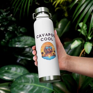 Peut inclure: Gourde isotherme blanche avec un couvercle et une base argentés. La gourde porte l'inscription "CAVAPOO COOL" au-dessus d'un dessin de chien Cavapoo portant des lunettes de soleil, sur une scène de plage au coucher du soleil.