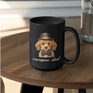 May include: Black ceramic mug featuring a Cavalier King Charles Spaniel and Poodle mix, wearing a top hat and bow tie. The text "cavapoo dad" is written in a script font below the dog illustration. The mug is on a wooden surface.