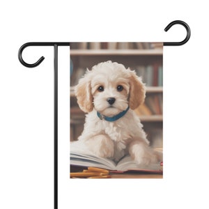 May include: A white and cream coloured puppy with brown eyes and a blue collar is sitting on a book with pencils on a table. The puppy is looking at the camera. The background is a bookshelf.