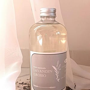 May include: A clear glass bottle with a silver cap, filled with a light-colored liquid. The label reads "Huile essentielle LAVANDIN GROSSO" with a lavender sprig illustration. The bottle sits on a bed of dried lavender buds.
