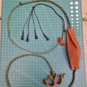 May include: A handmade slingshot and a loop of rope, both in shades of green, are displayed against a grid background. The slingshot features a brown leather pouch with a decorative pattern. The rope has a dark green braided design.