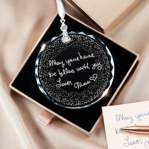 May include: A round, black crystal ornament with a handwritten message, "May your home be filled with joy, Love, Mom," is displayed in an open gift box. A matching note and pen are next to the box. The ornament hangs from a white ribbon.