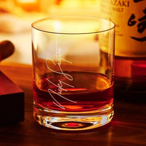 May include: A clear glass rocks glass with a personalized inscription that reads "Mickey Santiago" filled with amber colored liquid. The glass is sitting on a wooden surface.