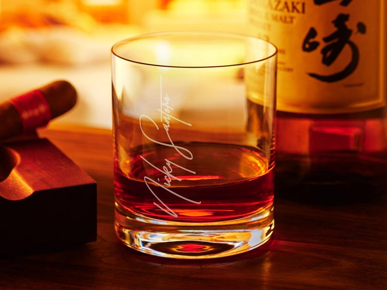 May include: A clear glass rocks glass with a personalized inscription that reads "Mickey Santiago" filled with amber colored liquid. The glass is sitting on a wooden surface.