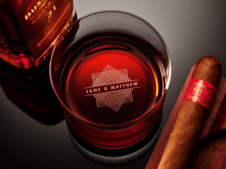 May include: A personalized glass with a red liquid inside. The glass has a geometric design and the text "JANE & MATTHEW" engraved on it. A brown cigar is resting on a wooden surface.