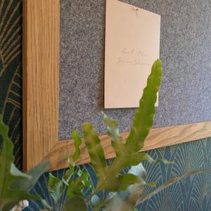 Puede incluir: Un tablero de anuncios rectangular de fieltro gris con un marco de madera de roble claro. Una tarjeta rectangular de color crema está sujeta al centro del tablero, con el texto manuscrito Let the Sun Shine en una fuente fina y clara.