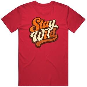 Puede incluir: Una camiseta roja con la frase "Stay Wild" en una fuente retro. El texto está en tonos de naranja, marrón y blanco, con un aspecto desgastado y vintage. La camiseta es de color rojo liso.