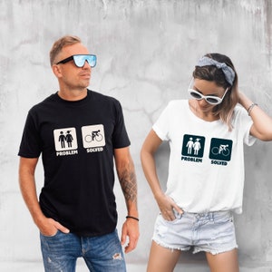 Può includere: Due magliette bianche con un design grafico di una coppia di figure stilizzate etichettata "problema" e una figura stilizzata che va in bicicletta etichettata "risolto".