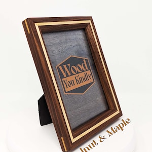 May include: A wooden picture frame with a walnut and maple wood inlay. The frame is standing on a white surface. The frame has a black backing and a clear glass front. The frame has a small stand on the back to prop it up. The frame has a logo on the front that says "Wood You Kindly".