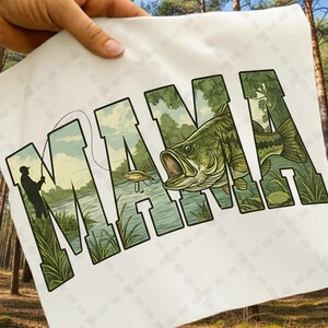 May include: White t-shirt featuring the word "MAMA" in large, green, stylized letters. The letters are filled with a fishing scene, including a fisherman, a fish, and a lake. The shirt is held up by a hand.
