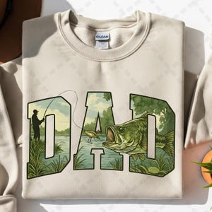 May include: Beige sweatshirt with the word "DAD" in a green and black design. The letters are filled with a fishing scene, including a fisherman, a fish, and a lake. The sweatshirt is made of a soft material.
