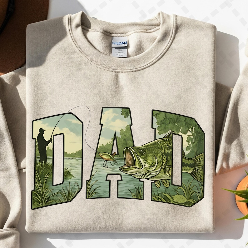 May include: Beige sweatshirt with the word "DAD" in a green and black design. The letters are filled with a fishing scene, including a fisherman, a fish, and a lake. The sweatshirt is made of a soft material.