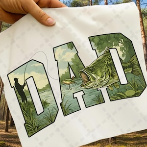May include: White fabric with the word "DAD" in green, with a fishing scene inside the letters. The letter "D" shows a fisherman casting a line, and the letter "D" shows a large mouth bass. The letter "A" shows a lure.
