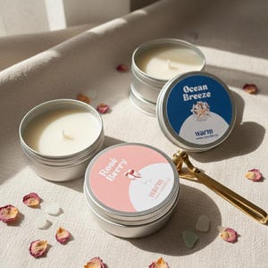 May include: A collection of small, round candles in silver tins. One tin is labelled "Ocean Breeze" and another "Rose Berry". Dried flower petals and small, white stones surround the candles. A gold-coloured tool is also visible.