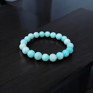 Op de afbeelding: Een lichtblauwe kralenarmband. De armband is gemaakt van ronde, gladde kralen in verschillende tinten lichtblauw. De armband ligt op een donkere ondergrond.