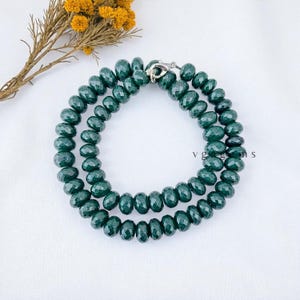 May include: A dark green beaded necklace with a silver clasp. The necklace is composed of multiple strands of faceted beads. The background is a white surface with a sprig of yellow flowers.