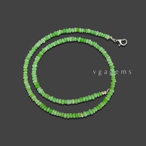 May include: A necklace composed of small, light green beads. The beads are strung together to form a necklace with a silver clasp. The necklace is displayed on a dark gray background. The text "vgagems" is visible.