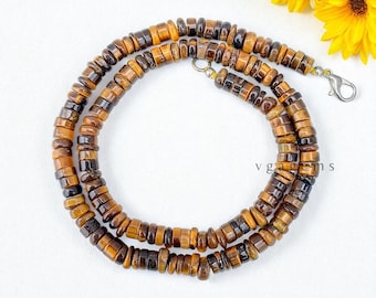 May include: A necklace made of small, round, tiger's eye beads in shades of brown and gold. The beads are strung together, forming a circular shape. The necklace has a silver clasp. The text "vgagems" is visible in the image.