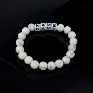 May include: White beaded bracelet with the word "NAME" spelled out in black letters on the beads.