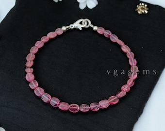 May include: A pink tourmaline gemstone bracelet with a silver clasp. The bracelet is made of oval-shaped beads and is displayed on a black fabric background.