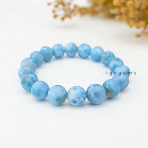 May include: A light blue beaded bracelet with a marbled effect. The round beads are strung together, forming a circular shape. The text "vgagems" is visible on the right side of the bracelet.