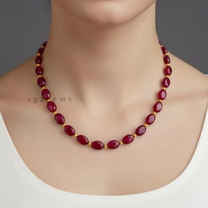 May include: A necklace featuring oval-shaped, deep red gemstones strung together with small gold-colored beads. The necklace is displayed against a neutral background, highlighting the rich color of the stones. The text "vga gems" is visible.