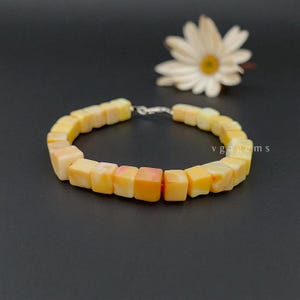 Pode incluir: Uma pulseira feita de contas quadradas amarelas e laranja. As contas são unidas por um fecho prateado. Uma margarida branca com centro amarelo está no fundo. O texto "vgagems" é visível à direita.
