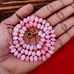May include: A pink opal gemstone necklace with a gold clasp. The necklace is made of small, round beads and is strung on a thin, pink thread.