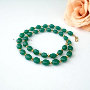 May include: A green beaded necklace with gold-colored accents. The necklace features oval-shaped green beads strung together with small gold-colored beads in between. The clasp is gold-colored. The text "vgagems" is visible on the left side.