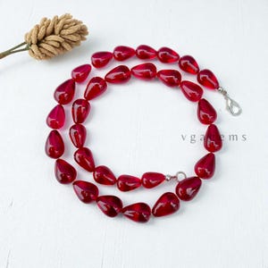 May include: A necklace featuring a strand of glossy, teardrop-shaped, deep red beads. The beads are strung together with a silver clasp. The background is a white surface with a decorative dried plant stem.