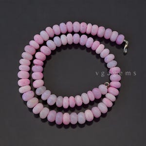 May include: A necklace composed of light pink, oval-shaped beads. The beads are strung together, forming a double strand. The necklace is displayed against a dark background. The text "vgagems" is visible.