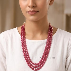 Puede incluir: Un collar de tres hilos con piedras preciosas ovaladas de color rojo intenso. El collar se lleva alrededor del cuello de una persona, con las piedras preciosas cayendo sobre el pecho. El collar está sobre una camiseta blanca. El texto "Vox GEMS" es visible.