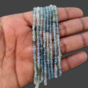 Puede incluir: Hebras de pequeñas cuentas de piedras preciosas de color azul claro y transparente. Las cuentas tienen una forma ligeramente irregular y están ensartadas. Las cuentas se sostienen en una mano. El texto "vgagems" es visible en la imagen.