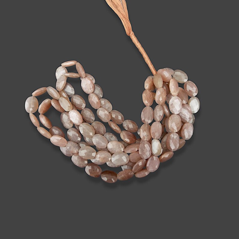 May include: A strand of oval-shaped sunstone beads in shades of peach and white. The beads are strung on a brown cord.