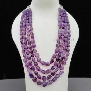 Può includere: Collana a più fili con perline di ametista ovali lucide in varie tonalità di viola. La collana è esposta su un busto di manichino bianco, che mostra il design a strati e le variazioni di colore delle pietre preziose.