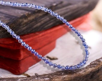 May include: A strand of blue gemstone beads, strung on a thin wire, is displayed on a wooden surface. The beads are small and round, with a slightly faceted appearance. The beads are a light blue color, with a hint of purple.