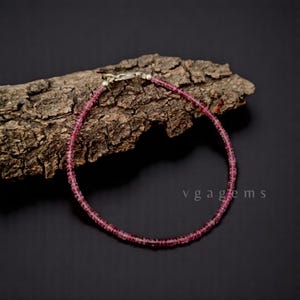 May include: A delicate pink gemstone bracelet with a silver clasp, resting on a piece of textured wood against a dark background. The bracelet is made of small, round beads. The text "vgagems" is visible in the lower right corner.