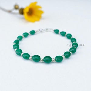 May include: A green beaded bracelet with silver accents. The bracelet features oval-shaped green beads strung together with small silver beads. The clasp is also silver. The background includes a yellow flower and the text "vgagems".