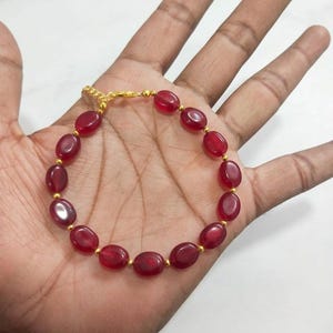May include: A bracelet featuring a series of smooth, oval-shaped deep red beads alternating with small, round gold-toned metal spacers. The jewelry piece includes a gold-toned metal chain and a lobster claw clasp closure for secure fastening.