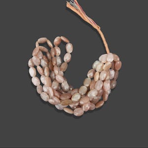 May include: A strand of oval-shaped sunstone beads in shades of peach and pink. The beads are strung on a thin cord and are ready to be used for jewellery making.