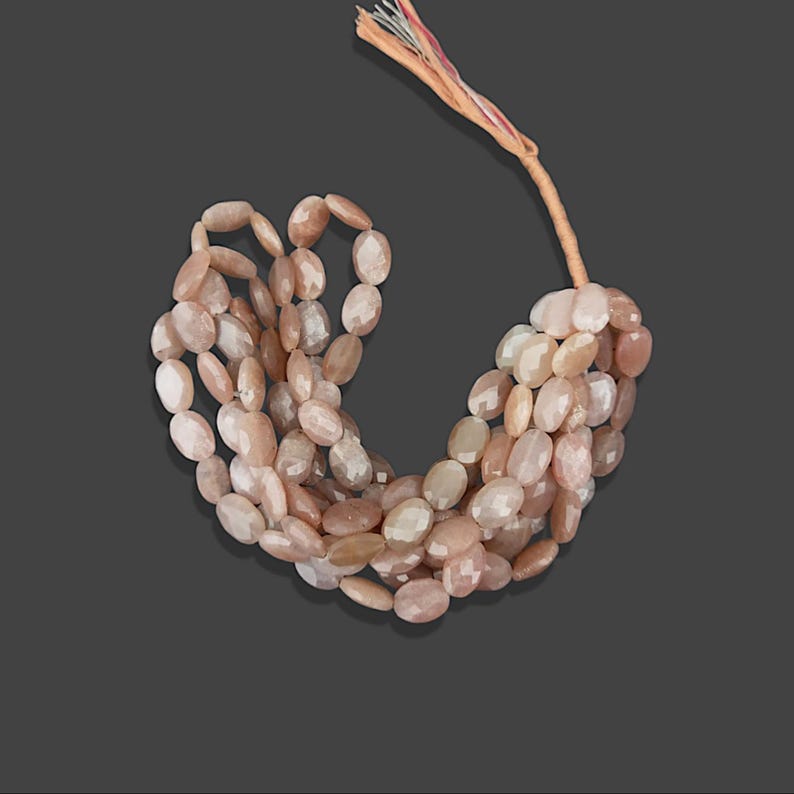 May include: A strand of oval-shaped sunstone beads in shades of peach and pink. The beads are strung on a thin cord and are ready to be used for jewellery making.